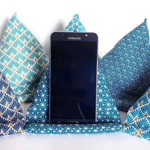 May include: A black Samsung smartphone resting in a triangular fabric stand. The stand features a teal and white geometric star pattern. Surrounding the central stand are four additional triangular fabric cushions in various blue and teal patterns.