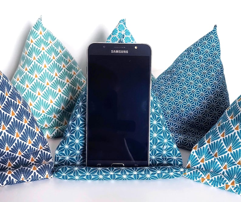 May include: A black Samsung smartphone resting in a triangular fabric stand. The stand features a teal and white geometric star pattern. Surrounding the central stand are four additional triangular fabric cushions in various blue and teal patterns.