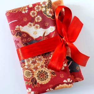 May include: A rectangular fabric bundle featuring a red base with a repeating pattern of white and gold floral blossoms and dark grey accents. The fabric is folded and secured with a bright red satin ribbon tied into a decorative bow on the side.