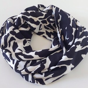 May include: A navy and white infinity scarf with a striking abstract design. The scarf is made from a soft, flowing material and is designed to be worn looped around the neck. The pattern features large, irregular shapes in a repeating design.