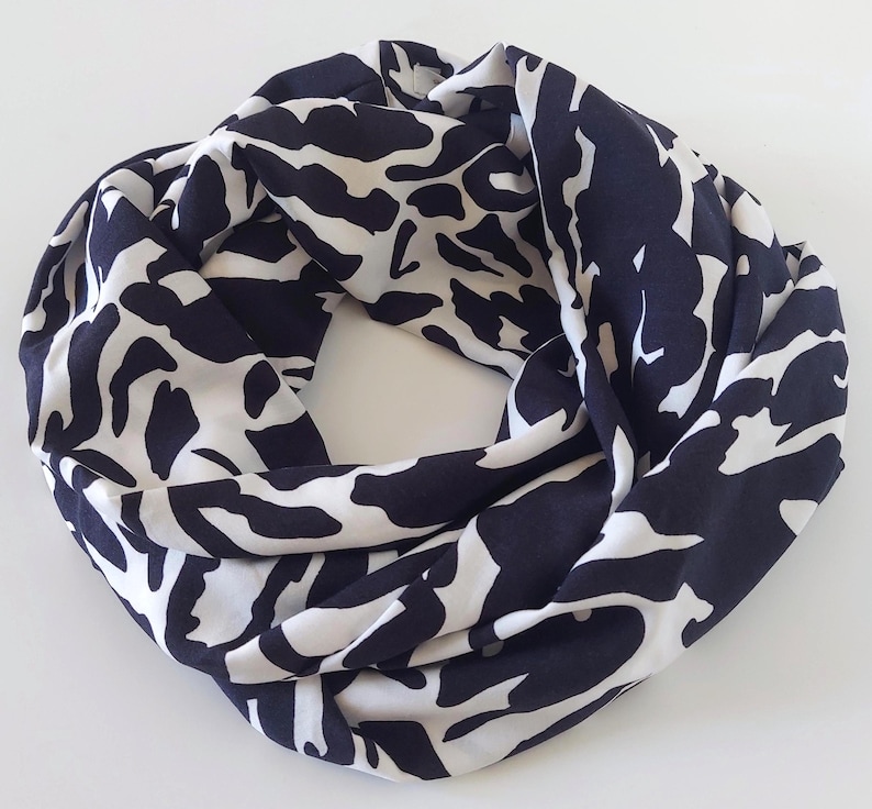 May include: A navy and white infinity scarf with a striking abstract design. The scarf is made from a soft, flowing material and is designed to be worn looped around the neck. The pattern features large, irregular shapes in a repeating design.
