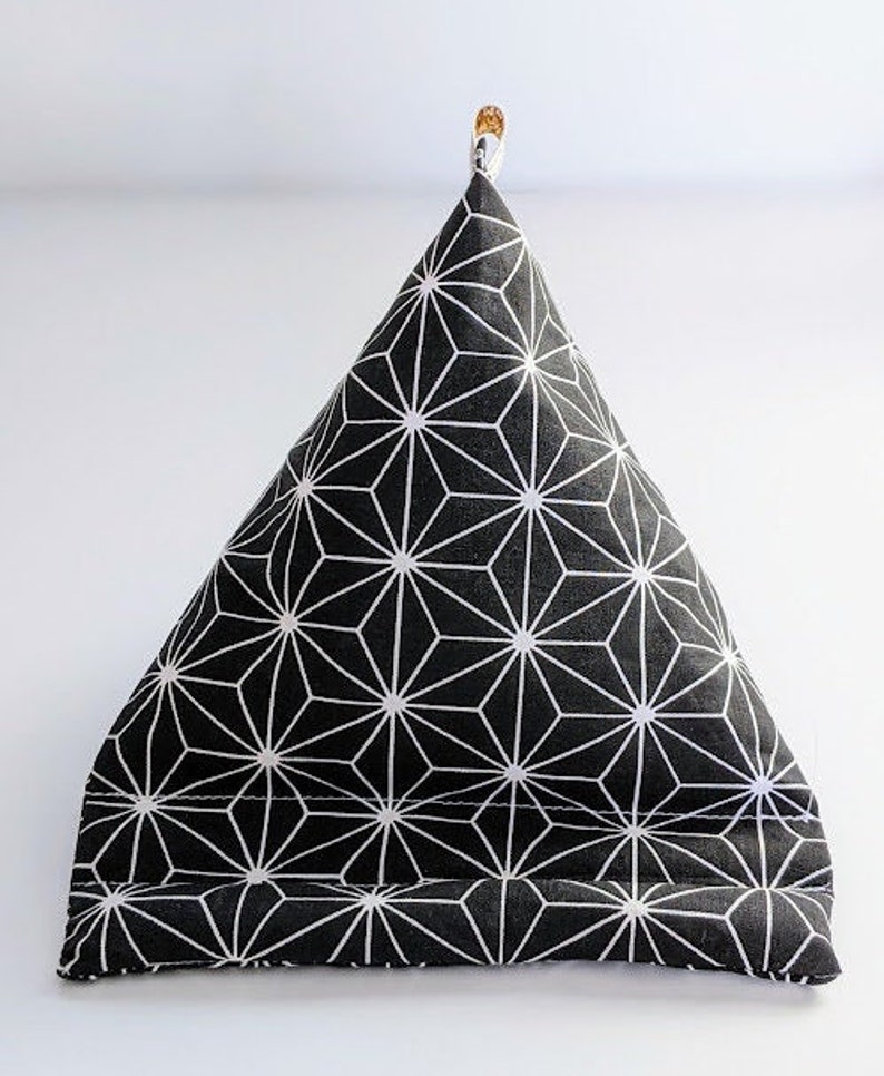 May include: A black and white geometric patterned triangular pillow. The pillow features a repeating starburst design in white against a black background. A small gold-colored clip is attached to the top of the pillow.