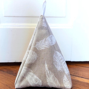 May include: A triangular doorstop made of tan fabric with a white feather pattern. It has a small white loop at the top for hanging. The doorstop is resting on a wooden floor.
