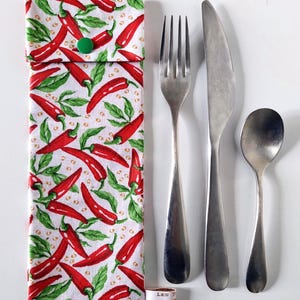 Puede incluir: Un estuche de tela blanca con un estampado de chiles rojos, un cierre de botón verde y un juego de cubiertos de acero inoxidable: tenedor, cuchillo y cuchara. También hay una pequeña etiqueta con texto.