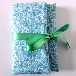 Può includere: Una custodia in tessuto rettangolare con un motivo floreale bianco e verde acqua. La custodia è piegata e fissata con un nastro di raso verde brillante legato a fiocco attorno al centro. Il tessuto ha una consistenza morbida con fiori.