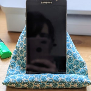 May include: A black Samsung smartphone resting in a triangular fabric stand. The stand features a teal and white geometric pattern with repeating hexagonal shapes. The phone screen is dark and reflective, showing a faint silhouette of the surroundings.
