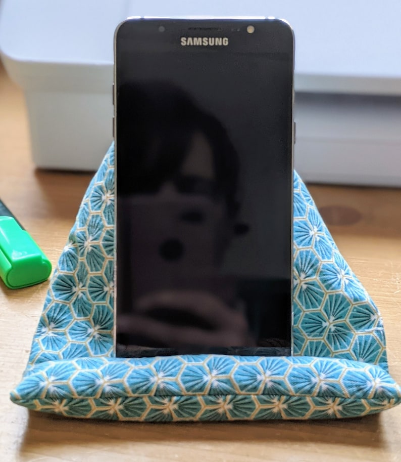 May include: A black Samsung smartphone resting in a triangular fabric stand. The stand features a teal and white geometric pattern with repeating hexagonal shapes. The phone screen is dark and reflective, showing a faint silhouette of the surroundings.