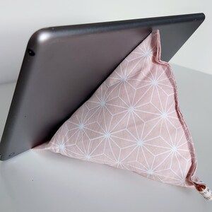 May include: A tablet propped up by a pink and white geometric patterned pillow stand. The pillow is triangular and made of fabric. The tablet is gray and angled for viewing.