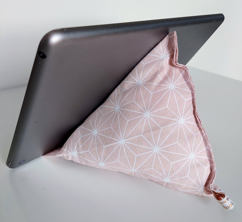 May include: A tablet propped up by a pink and white geometric patterned pillow stand. The pillow is triangular and made of fabric. The tablet is gray and angled for viewing.