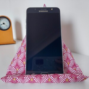 May include: A Samsung smartphone rests on a pink and white patterned fabric stand. The stand is triangular, with a fan-like design in white and gold. A small, orange clock is visible in the background.