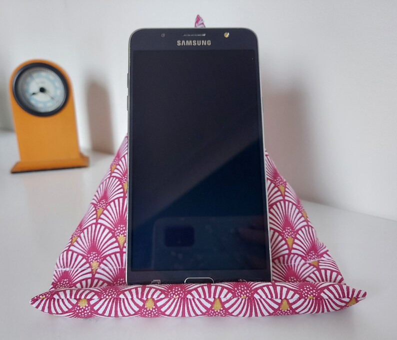 May include: A Samsung smartphone rests on a pink and white patterned fabric stand. The stand is triangular, with a fan-like design in white and gold. A small, orange clock is visible in the background.