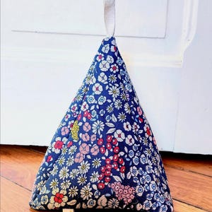 May include: A triangular doorstop in blue floral fabric. The fabric is patterned with small white, pink, and red flowers. A small tag is attached to the bottom. A white fabric loop is attached to the top for hanging.