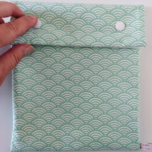 May include: A square fabric pouch featuring a repeating mint green and white wave pattern. The pouch has a fold-over flap secured by two white plastic snap buttons. A small white fabric tag with partial text is visible on the bottom right corner.