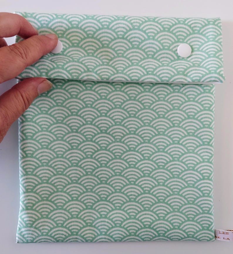 May include: A square fabric pouch featuring a repeating mint green and white wave pattern. The pouch has a fold-over flap secured by two white plastic snap buttons. A small white fabric tag with partial text is visible on the bottom right corner.