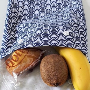 May include: A fabric snack pouch featuring a blue and white Japanese wave pattern. The pouch is secured with two white plastic snap buttons and contains a wrapped madeleine cake, a brown fuzzy kiwi fruit, and a yellow banana inside the opening.