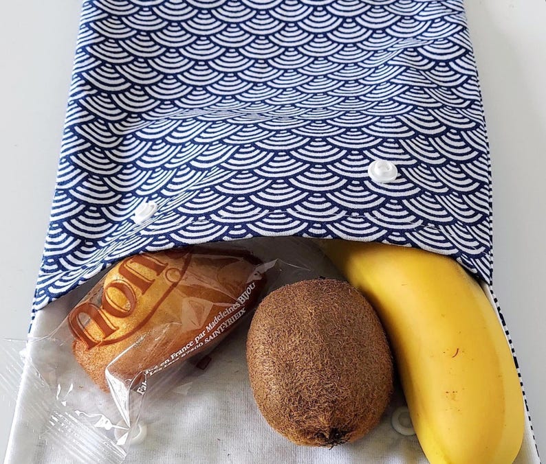 May include: A fabric snack pouch featuring a blue and white Japanese wave pattern. The pouch is secured with two white plastic snap buttons and contains a wrapped madeleine cake, a brown fuzzy kiwi fruit, and a yellow banana inside the opening.