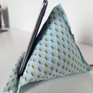 May include: A light blue fabric phone stand with a fan-like pattern in gold and white, holding a silver smartphone. The stand is triangular and made of soft fabric.