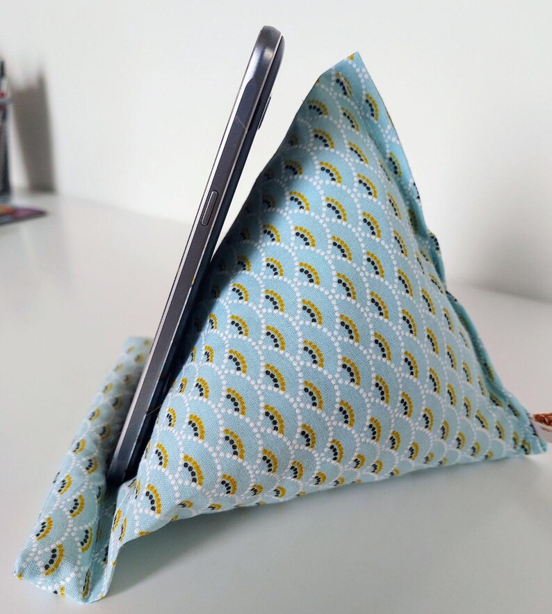 May include: A light blue fabric phone stand with a fan-like pattern in gold and white, holding a silver smartphone. The stand is triangular and made of soft fabric.