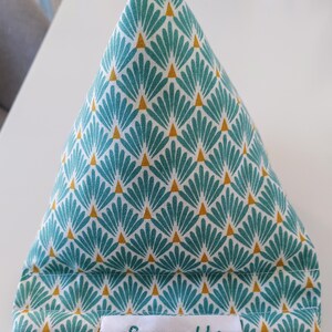 May include: A triangular fabric tablet or phone stand featuring a teal and white fan-shaped pattern with small gold triangles. A white rectangular fabric label is stitched to the bottom front with the word Soeurette embroidered in teal thread.