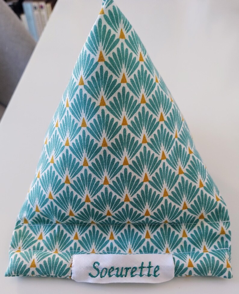 May include: A triangular fabric tablet or phone stand featuring a teal and white fan-shaped pattern with small gold triangles. A white rectangular fabric label is stitched to the bottom front with the word Soeurette embroidered in teal thread.