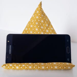 May include: A yellow and white patterned fabric phone stand with a triangular back support and a rectangular base. A black Samsung smartphone rests on the stand. The fabric design features a repeating floral pattern.