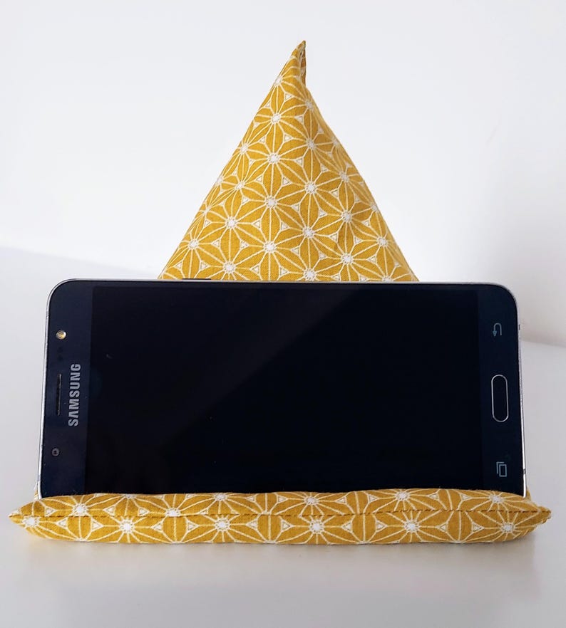 May include: A yellow and white patterned fabric phone stand with a triangular back support and a rectangular base. A black Samsung smartphone rests on the stand. The fabric design features a repeating floral pattern.