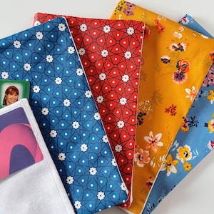 Può includere: Una collezione di sacchetti di stoffa colorati con vari motivi. I sacchetti includono disegni in blu con stelle bianche, rosso con forme geometriche bianche, giallo senape con stampe floreali e azzurro con stampe floreali. Un sacchetto bianco contiene una carta.