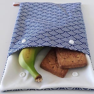 May include: A rectangular fabric snack pouch featuring a blue and white repeating fan-shaped pattern. The pouch is open, revealing a white interior lining, a single yellow banana, and two rectangular brown cookies. It has white plastic snap buttons.