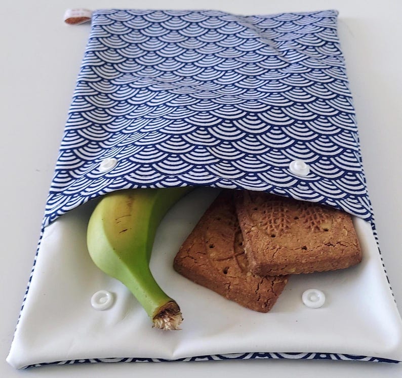 May include: A rectangular fabric snack pouch featuring a blue and white repeating fan-shaped pattern. The pouch is open, revealing a white interior lining, a single yellow banana, and two rectangular brown cookies. It has white plastic snap buttons.