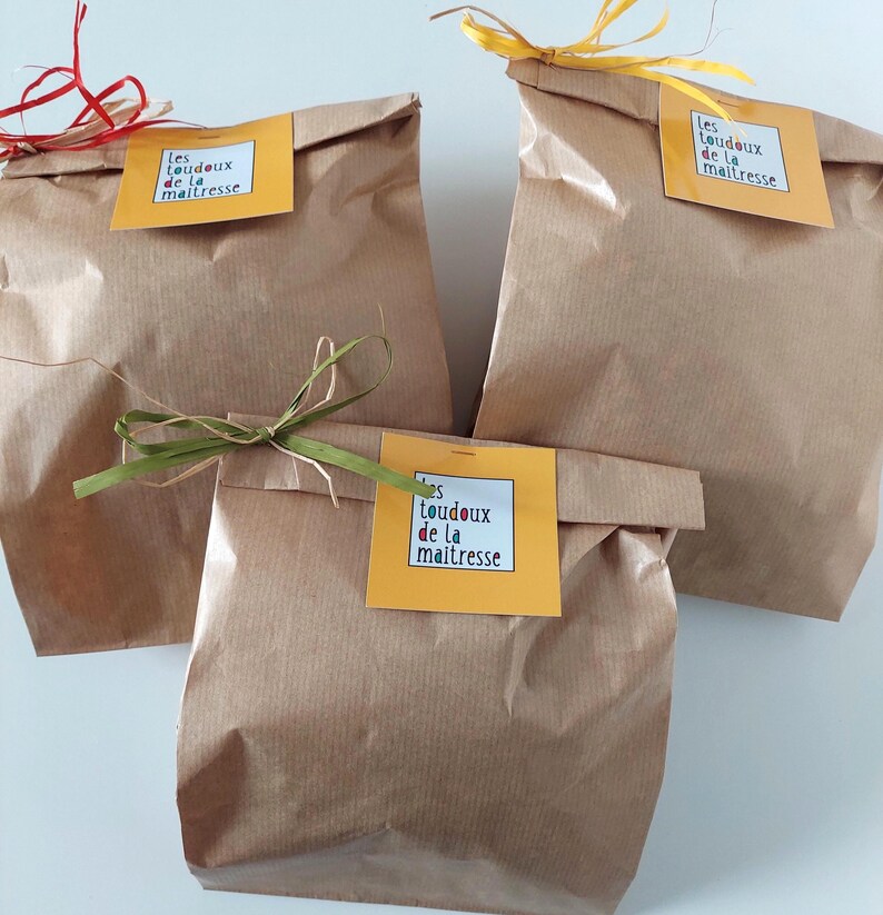 May include: Three brown paper bags with folded tops, each adorned with a yellow tag featuring text. The bags are tied with ribbons in red, yellow, and green. The text on the tags reads "les toudoux de la maitresse."