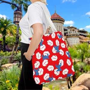 A person is standing in front of a tropical landscape with palm trees and a castle-like structure in the background. The person is carrying a red tote bag with a leaf pattern on it.