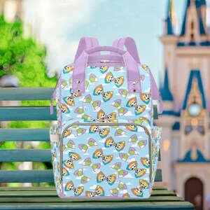 A colorful patterned backpack with cartoon animal designs is displayed on a wooden bench in front of a castle-like building.