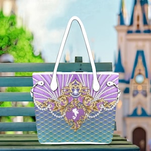 A handbag with a purple and gold pattern, featuring a mermaid design, is displayed on a bench in front of a castle.