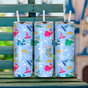 May include: Three stainless steel tumblers with white straws. Each tumbler features a light blue background with a repeating pattern of fairies, witch hats, and stars. The tumblers are designed for cold beverages.