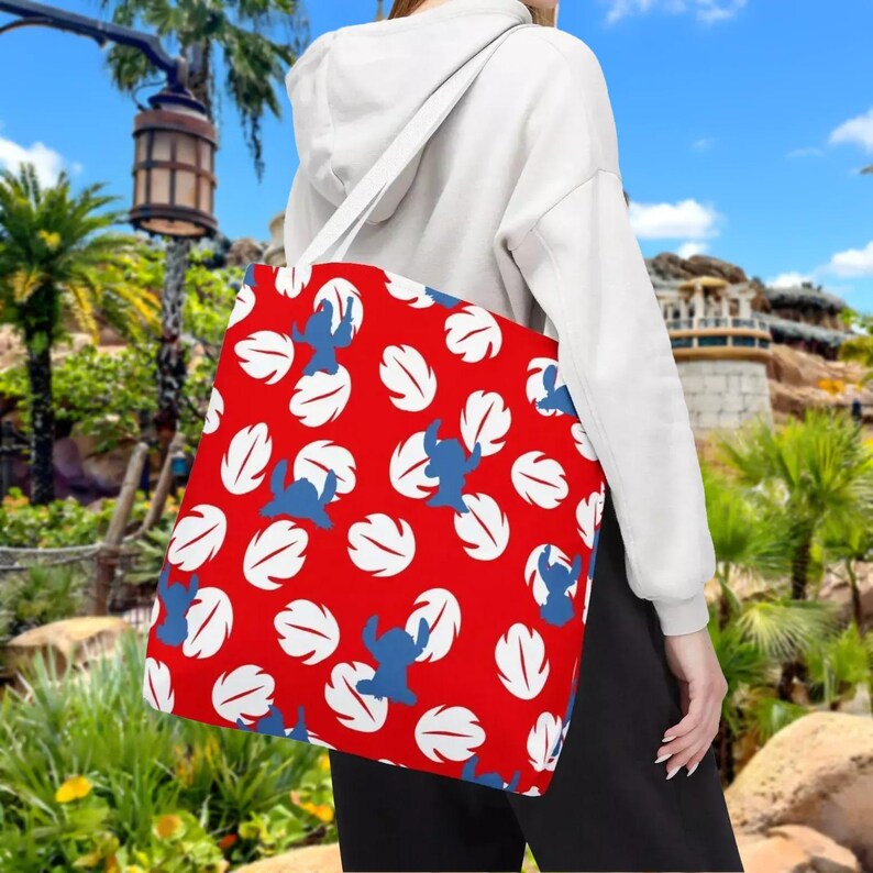 A person is standing in front of a tropical landscape, wearing a white hoodie and carrying a red tote bag with a pattern of white and blue leaves.