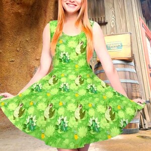 May include: A sleeveless green dress featuring an all-over repeating pattern of cartoon alligators and light green water lilies. The dress has a round neckline and a flared skirt silhouette with a vibrant, multi-tonal green color palette.