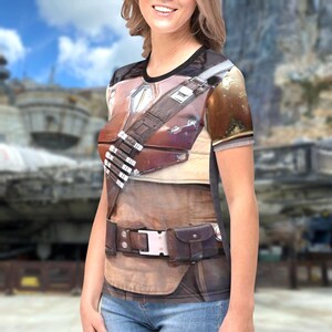 A woman wearing a t-shirt with a detailed graphic design of a leather belt and gun holster, standing in front of a blurred background.