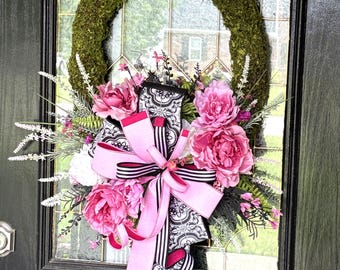 Op de afbeelding: Een decoratieve krans met een met mos bedekte basis, versierd met roze pioenrozen, witte bloemen en groen. Een grote roze strik met zwart-wit patroonlint zorgt voor een opvallend contrast. De krans is ontworpen voor deurdecoratie.
