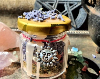 May include: A small glass jar with a gold lid, filled with dried herbs and lavender. A silver sun charm hangs from the jar. The jar is surrounded by crystals and a small cauldron, suggesting a mystical theme.
