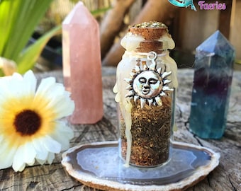 May include: A small glass bottle filled with dried herbs and topped with a cork stopper. The bottle is decorated with a silver charm of a sun with a face. The bottle is sitting on a white agate slice.