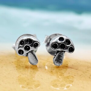 May include: A pair of silver-toned stud earrings shaped like mushrooms. Each earring features a rounded cap with five recessed black circular details and a textured stem. The metal has a polished finish with darkened accents in the crevices.