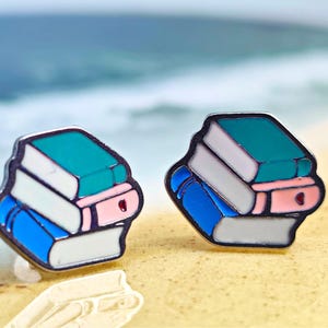 Puede incluir: Un par de pendientes de esmalte con una pila de libros. Cada libro es azul, blanco, turquesa y rosa. Los pendientes tienen un contorno plateado. El fondo es una playa.