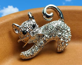 May include: A silver-toned cat brooch featuring a polished metal finish and encrusted with numerous clear, sparkling rhinestones. The cat is depicted in a walking pose with a curled tail, pointed ears, and two small black stones for eyes.