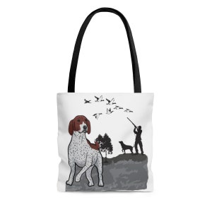 May include: White tote bag with a brown and white spotted dog, a hunter with a gun, and a flock of birds flying overhead. The dog is looking to the right, and the hunter is aiming his gun at the birds. The background is a gray and white landscape.