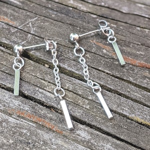 May include: A pair of silver dangle earrings with a small ball at the top and a chain that leads to a small rectangular bar. The earrings are on a wooden surface.