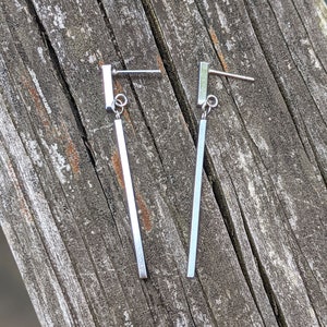 May include: A pair of silver dangle earrings with a long, thin bar pendant. The earrings are made of metal and have a simple, minimalist design.