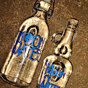 May include: Two clear glass bottles with cork stoppers and blue labels that say "Moon Water". The bottles are sitting on a brown textured surface.