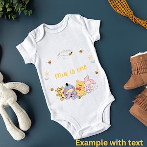 May include: White baby onesie with a Winnie the Pooh theme. The onesie has a yellow and white floral design with bees and the text "Mia is one". There are four Winnie the Pooh characters on the onesie: Winnie the Pooh, Piglet, Tigger, and Eeyore.