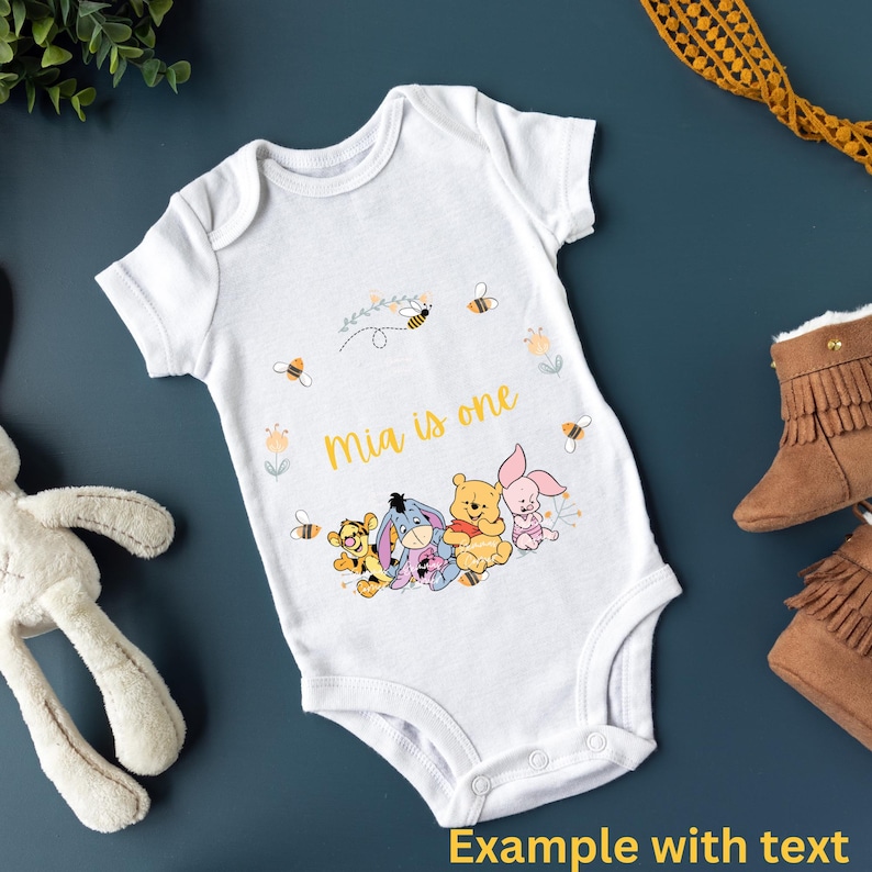 May include: White baby onesie with a Winnie the Pooh theme. The onesie has a yellow and white floral design with bees and the text "Mia is one". There are four Winnie the Pooh characters on the onesie: Winnie the Pooh, Piglet, Tigger, and Eeyore.