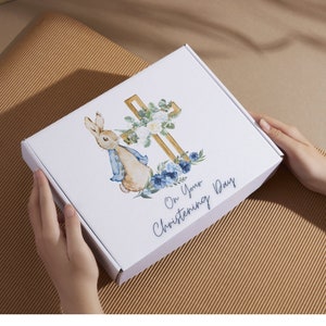 Peut inclure: Une bo&icirc;te cadeau blanche avec une illustration aquarelle bleue et blanche d'un lapin et d'une croix dor&eacute;e. Le texte "On Your Christening Day" est imprim&eacute; sur la bo&icirc;te.
